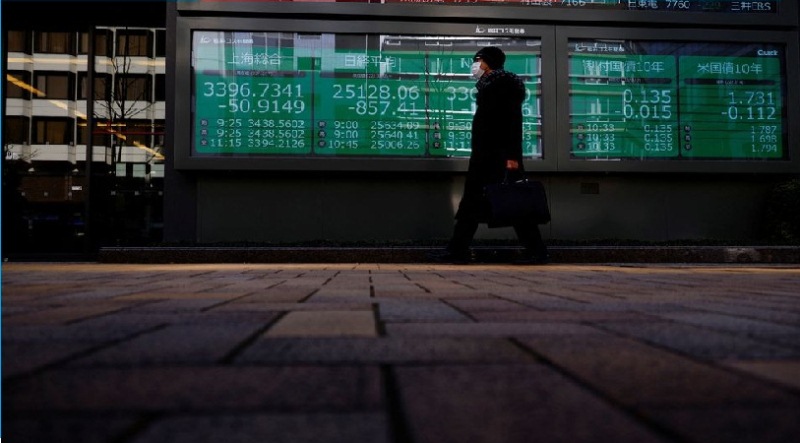 Seorang pria yang mengenakan masker pelindung, di tengah wabah penyakit virus corona (COVID-19), berjalan melewati papan elektronik yang menampilkan indeks Shanghai Composite, indeks Nikkei, dan Dow Jones Industrial Average di luar sebuah pialang di Tokyo